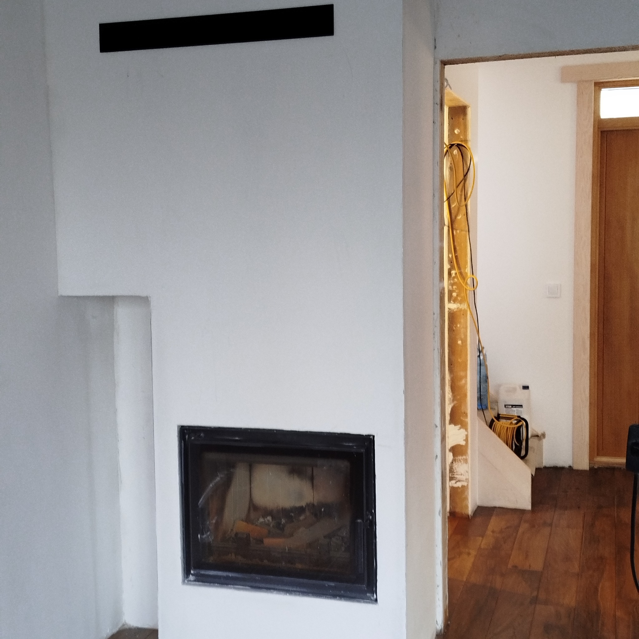 Biała ściana z czarnym wkładem kominkowym i elementem dekoracyjnym nad nim. Widoczny fragment podłogi i przejście do innego pomieszczenia w trakcie prac.