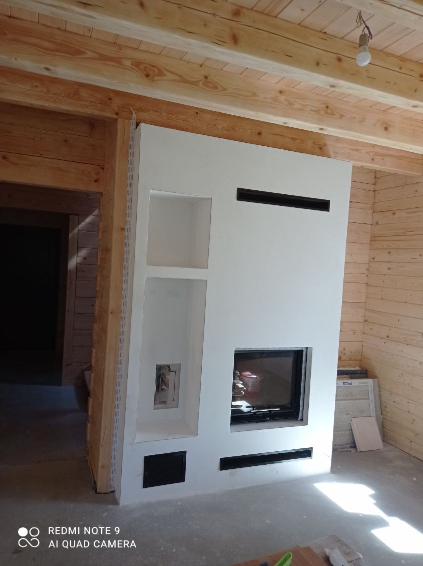 Biały kominek z wkładem i półkami w surowym wnętrzu z drewnianymi ścianami i sufitem. Widoczne kratki wentylacyjne i instalacja elektryczna.