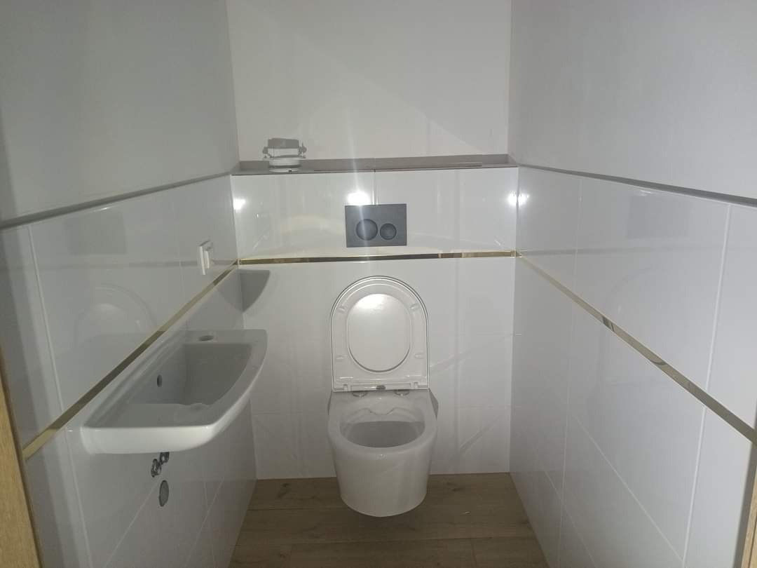 Biała łazienka z umywalką i toaletą w minimalistycznym stylu, wykończona białymi płytkami z delikatnymi złotymi akcentami. Nowoczesne wnętrze.