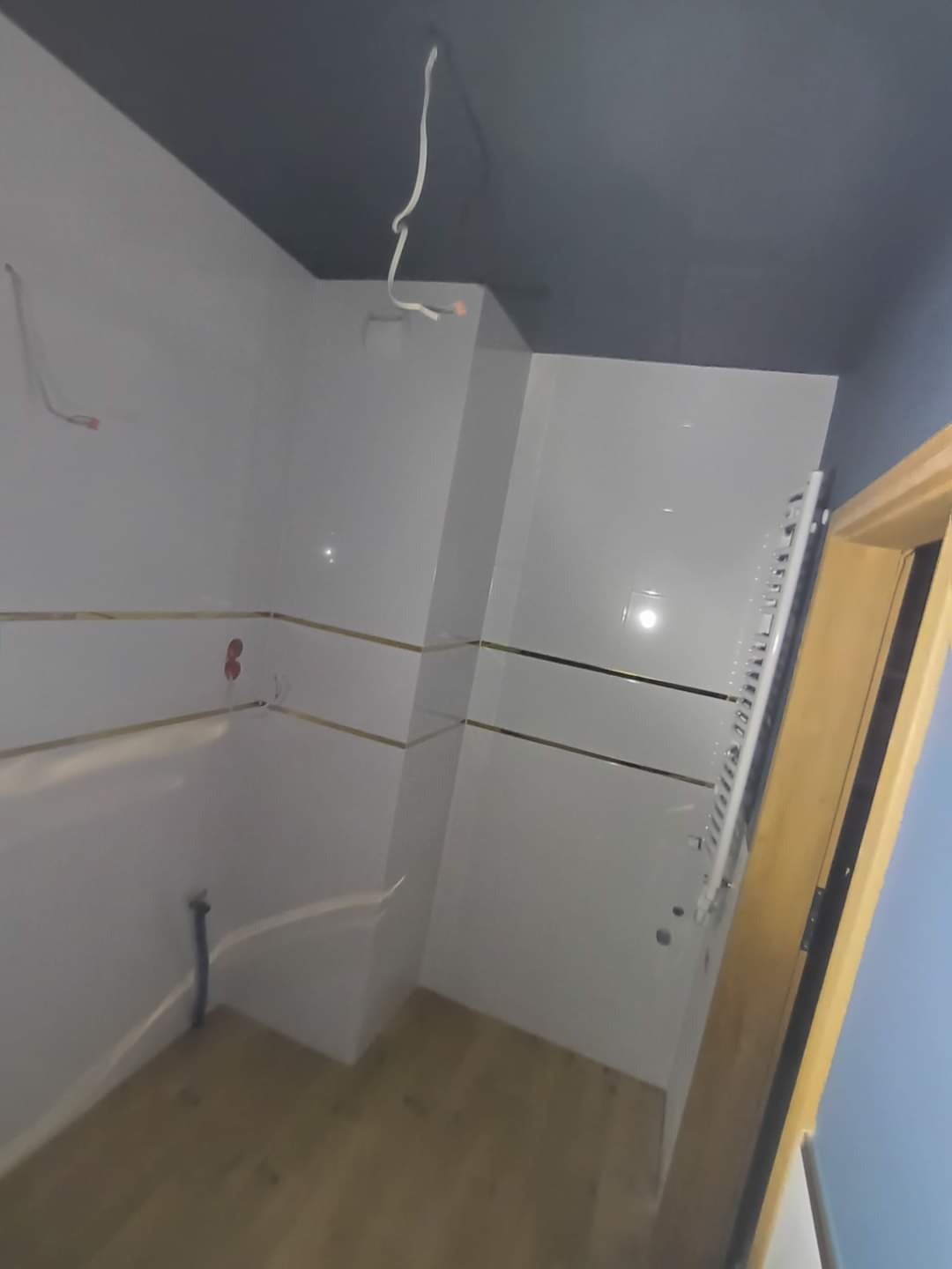 Biała łazienka z złotymi akcentami i grzejnikiem drabinkowym. Ciemny sufit i drewniane drzwi dodają kontrastu. Widoczne niedokończone prace elektryczne.