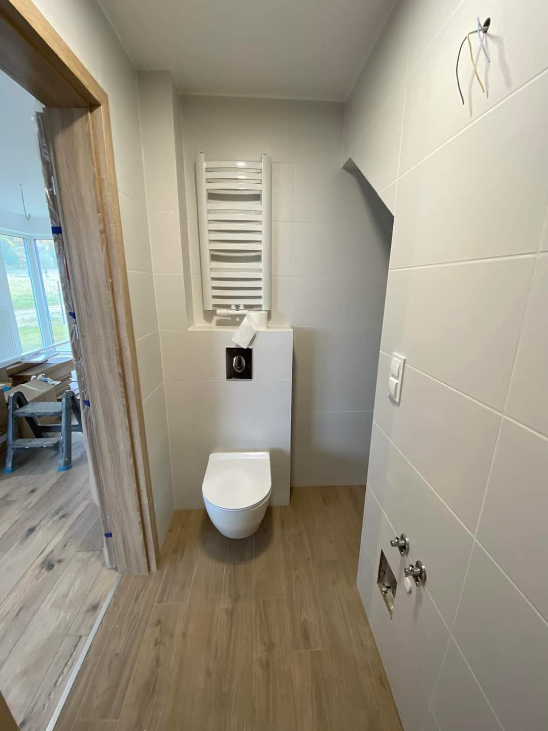Biała, nowoczesna łazienka z drewnopodobnymi płytkami podłogowymi. Widoczna podwieszana toaleta, grzejnik drabinkowy i częściowo wykończone przyłącza wodne.