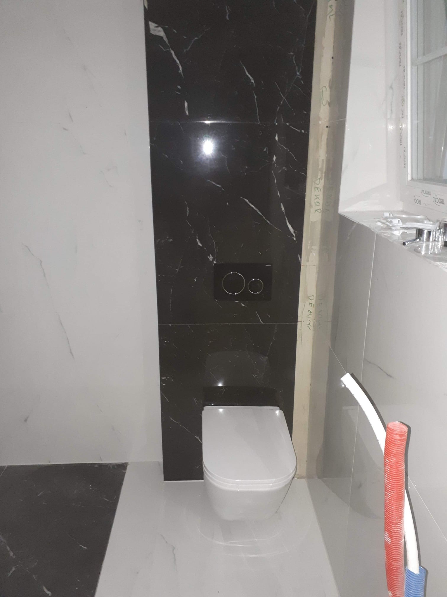 Nowoczesna łazienka w trakcie wykończenia: biała ceramika sanitarna na tle czarnych płytek imitujących marmur. Widoczne rury instalacyjne i fragment okna.