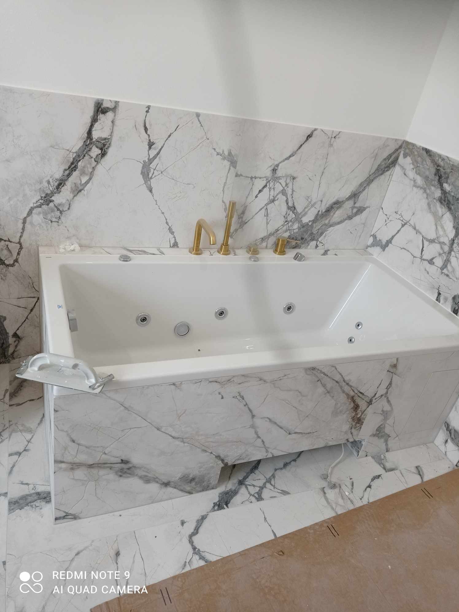 Biała wanna z hydromasażem, obłożona płytkami imitującymi marmur, z widoczną pacą glazurniczą, złota armatura, wnętrze łazienki w trakcie wykończenia.
