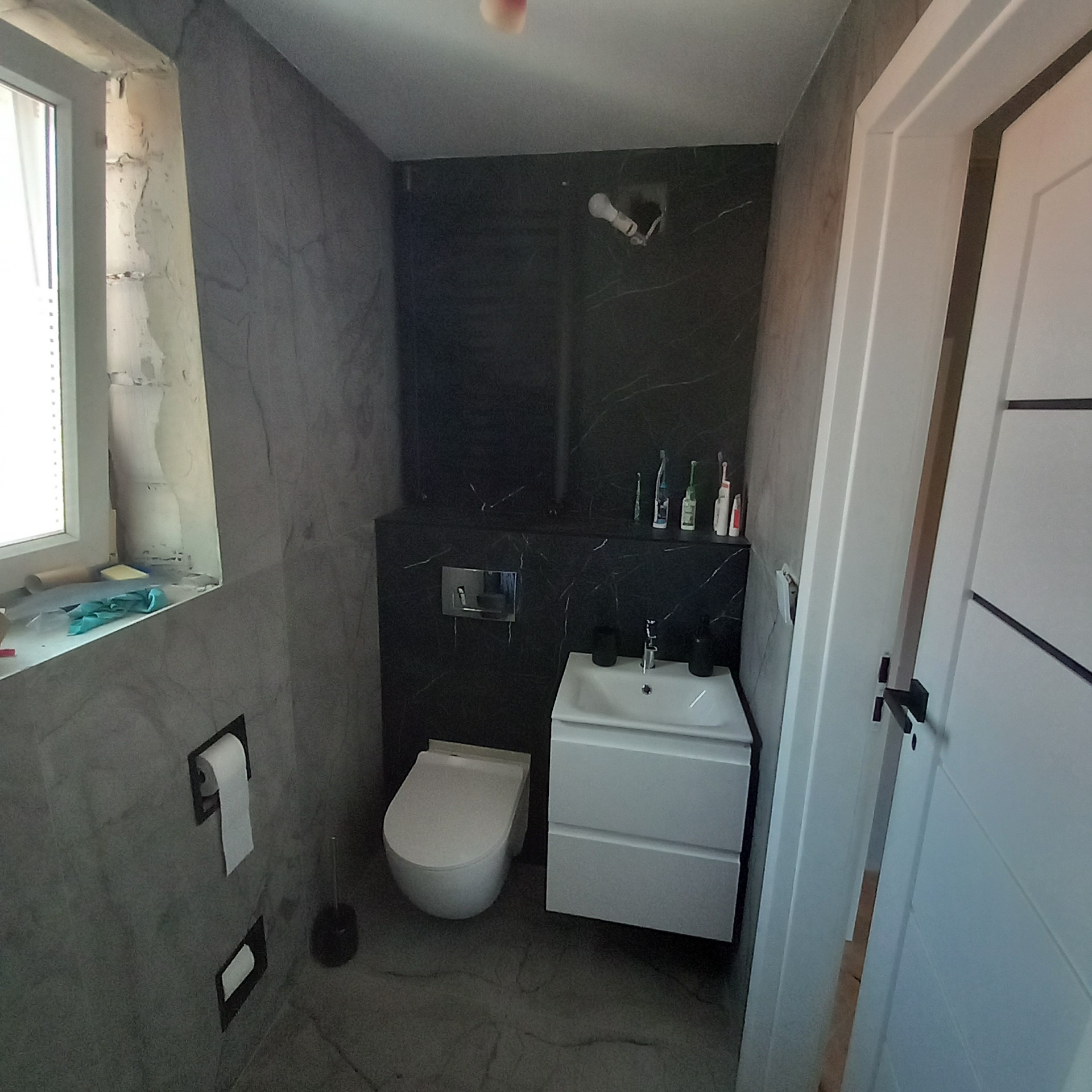 Nowoczesna łazienka po remoncie: szare płytki imitujące marmur, wisząca toaleta, biała szafka z umywalką i czarne akcenty. Okno w trakcie wykończenia.