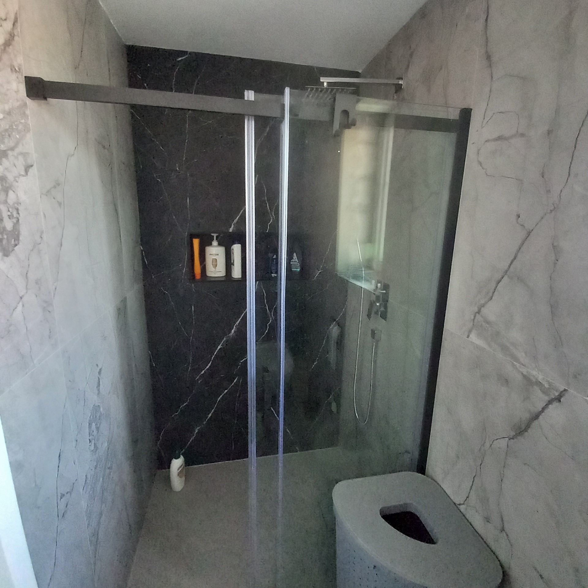 Nowoczesna kabina prysznicowa z przesuwnymi drzwiami, wykończona płytkami imitującymi marmur w odcieniach szarości i czerni, z wnęką na kosmetyki.