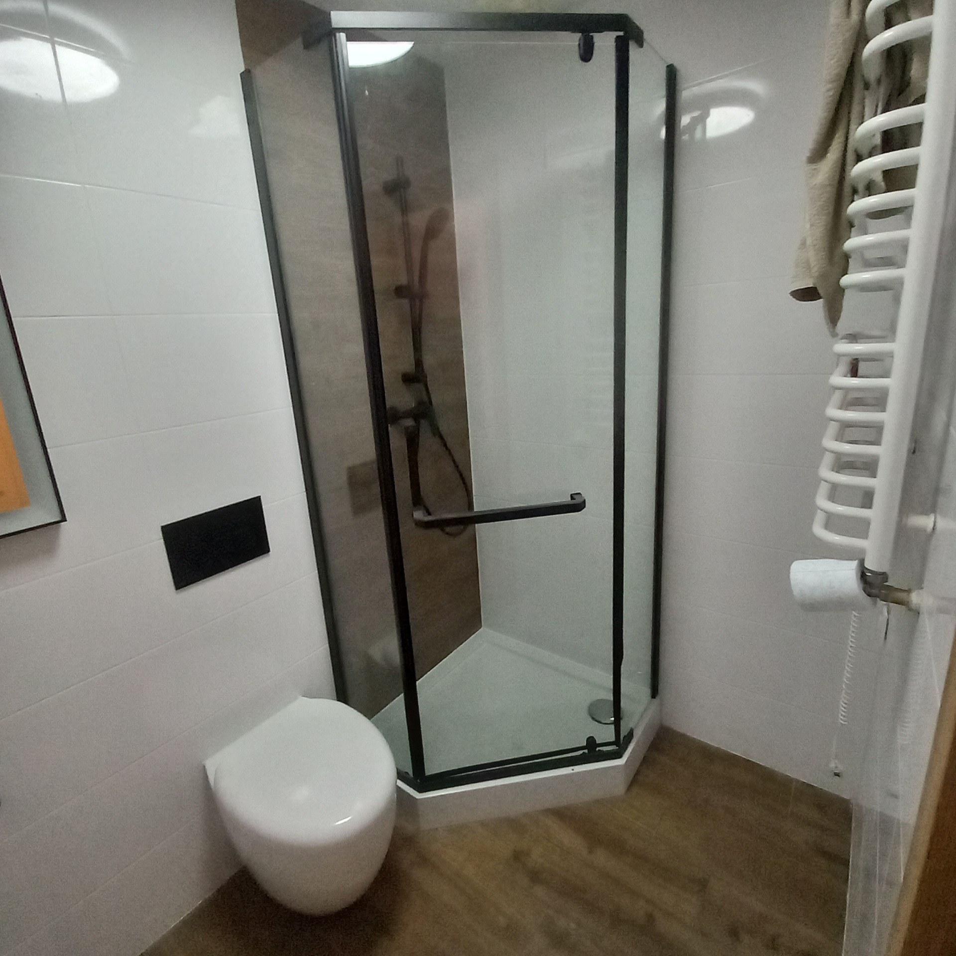 Nowoczesna kabina prysznicowa z czarnymi profilami w wykończonej łazience. Białe płytki na ścianach, drewnopodobne panele na podłodze. Widoczny grzejnik drabinkowy.