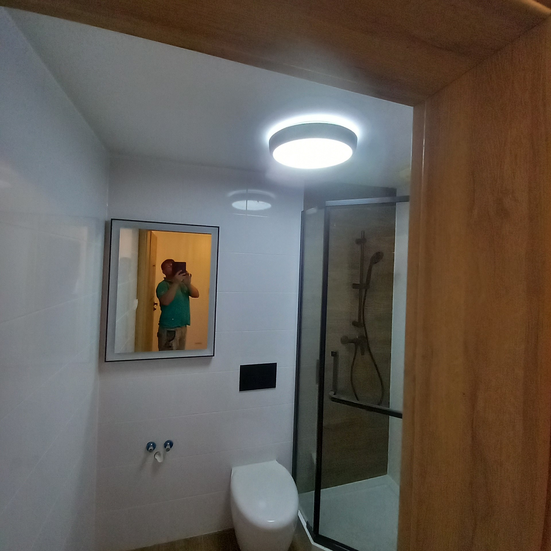 Nowoczesna, jasna łazienka z kabiną prysznicową i podświetlanym lustrem. Białe płytki na ścianach, minimalistyczna toaleta, ciemna rama kabiny.