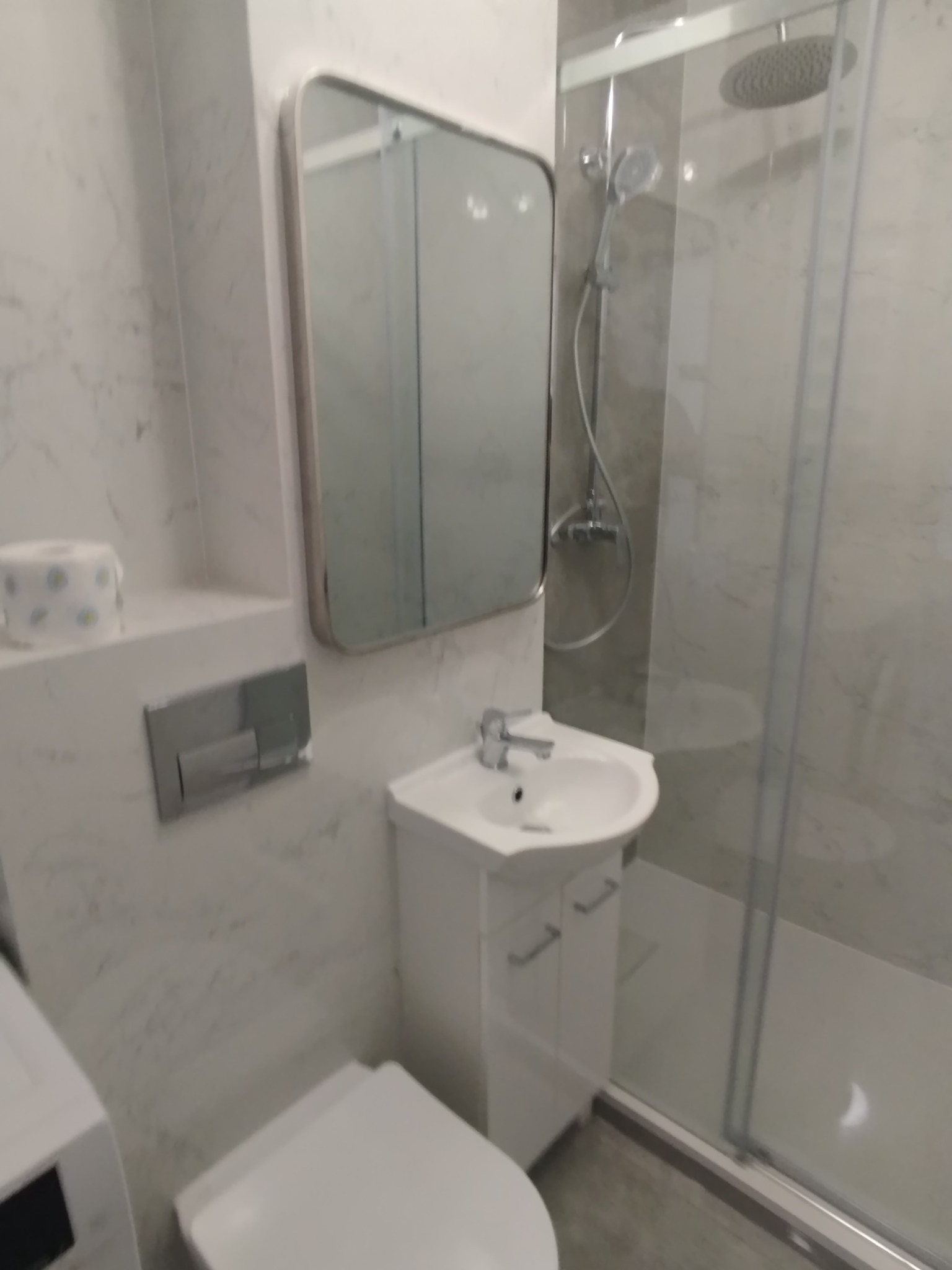 Elegancka, jasna łazienka z kabiną prysznicową typu walk-in, umywalką z szafką i toaletą. Marmurowe płytki ścienne, lustro w metalowej ramie. Minimalistyczny design.