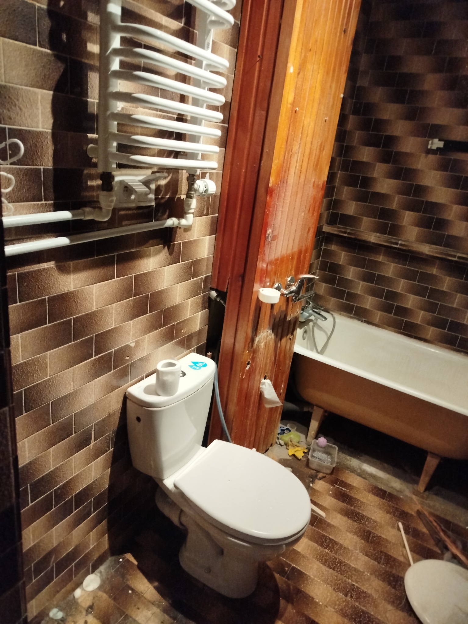 Brązowe płytki w łazience z wanną, toaletą i grzejnikiem drabinkowym. Widoczne ślady po remoncie. Ujęcie z bliska, niskie oświetlenie.