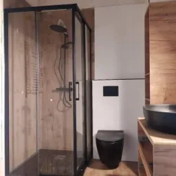 Nowoczesna łazienka z czarną kabiną prysznicową, wiszącą toaletą i umywalką na blacie z drewnopodobnymi szafkami, wykończona płytkami imitującymi drewno i białymi ścianami.