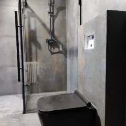 Nowoczesna łazienka z czarną podwieszaną toaletą, kabiną prysznicową ze stali i szklanymi drzwiami, oraz płytkami imitującymi rdzę na ścianach.