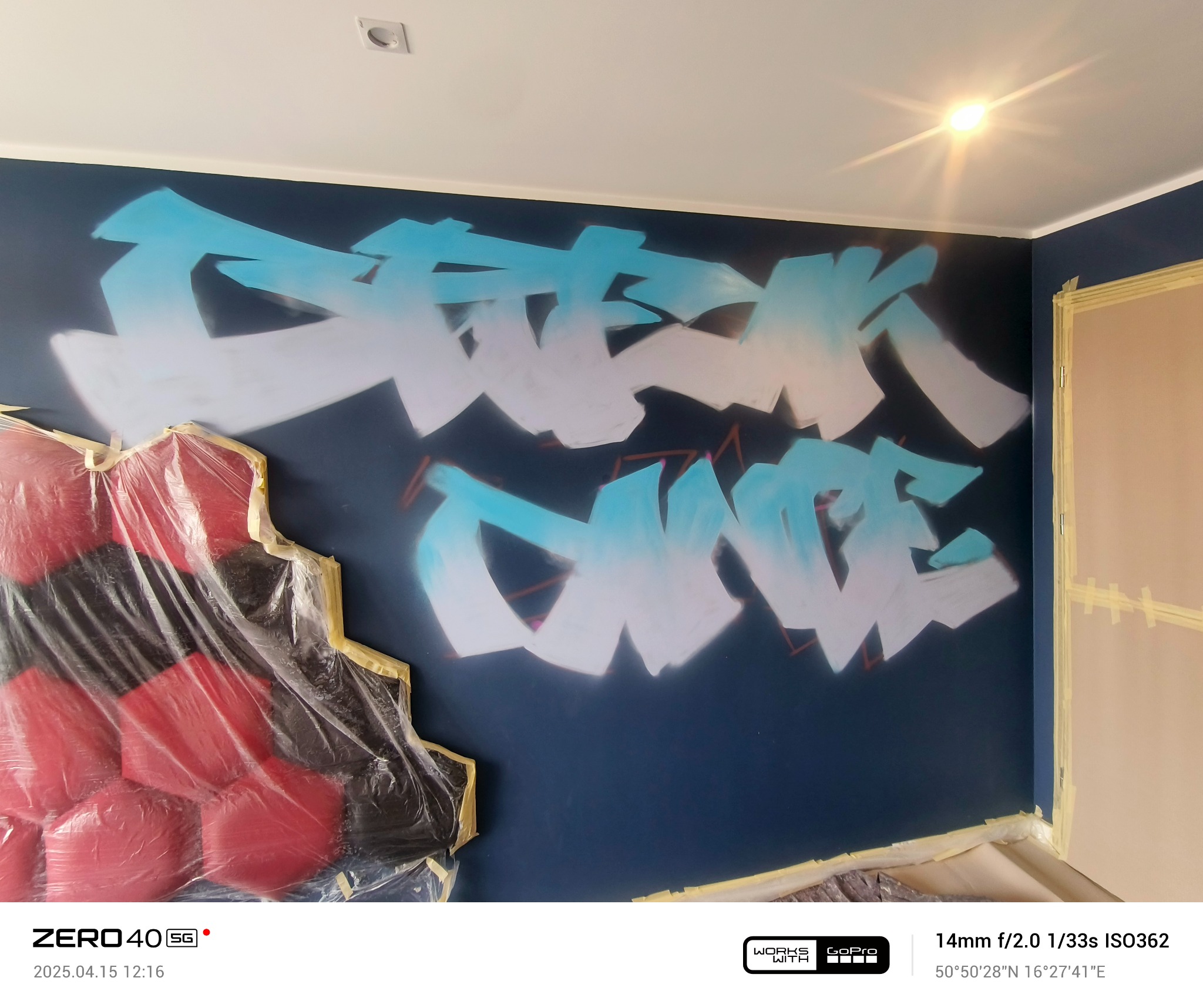 Niebiesko-biały graffiti napis na ciemnej ścianie, zabezpieczony fragment ściany i narożnik drzwi oklejone taśmą malarską, podłoga zabezpieczona folią.