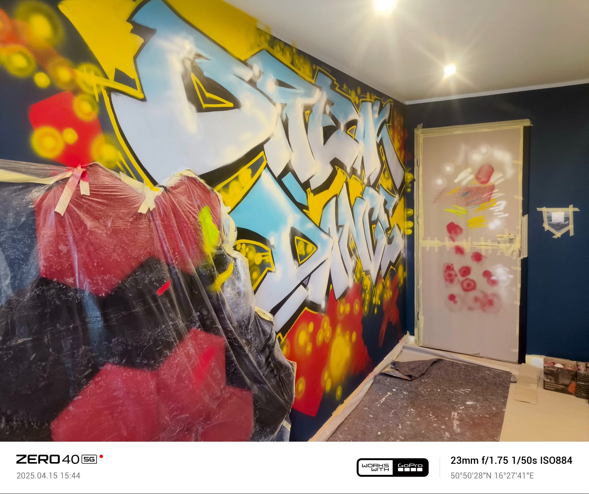 Graffiti na ścianie w trakcie tworzenia, częściowo zakryte folią ochronną, zabezpieczone drzwi w tle, widoczne ślady farby na podłodze.