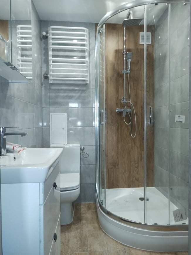 Nowoczesna łazienka z kabiną prysznicową narożną, grzejnikiem drabinkowym i umywalką z szafką, wykończona płytkami w odcieniach szarości i brązu.