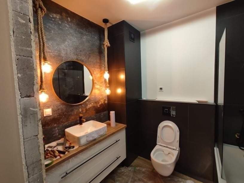 Nowoczesna łazienka w trakcie wykańczania: surowa ściana z cegły kontrastuje z dekoracyjną ścianą z efektem rdzy, okrągłe lustro z podświetleniem na sznurach, biała umywalka nablatowa na szafce...