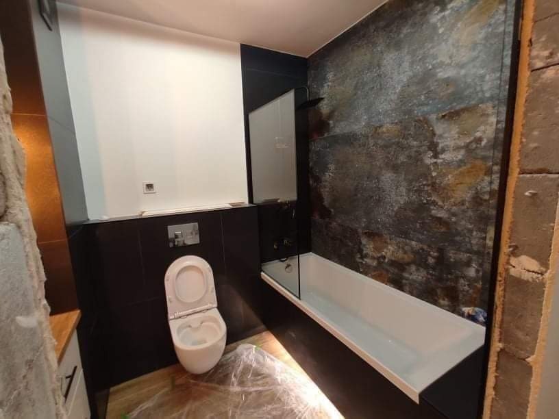 Nowoczesna łazienka w trakcie wykańczania, z białą wiszącą toaletą, wanną i ścianą z płytek imitujących kamień, zabezpieczona folią podłoga.