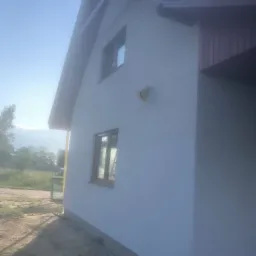 Boczny widok domu z jasną, świeżo pomalowaną elewacją; widoczne dwa okna z brązowymi ramami, fragment dachu z szarymi dachówkami i element instalacji na ścianie.