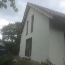 Świeżo otynkowana elewacja domu jednorodzinnego z ciemną dach&oacute;wką, widok z boku, w tle drzewa i pochmurne niebo.