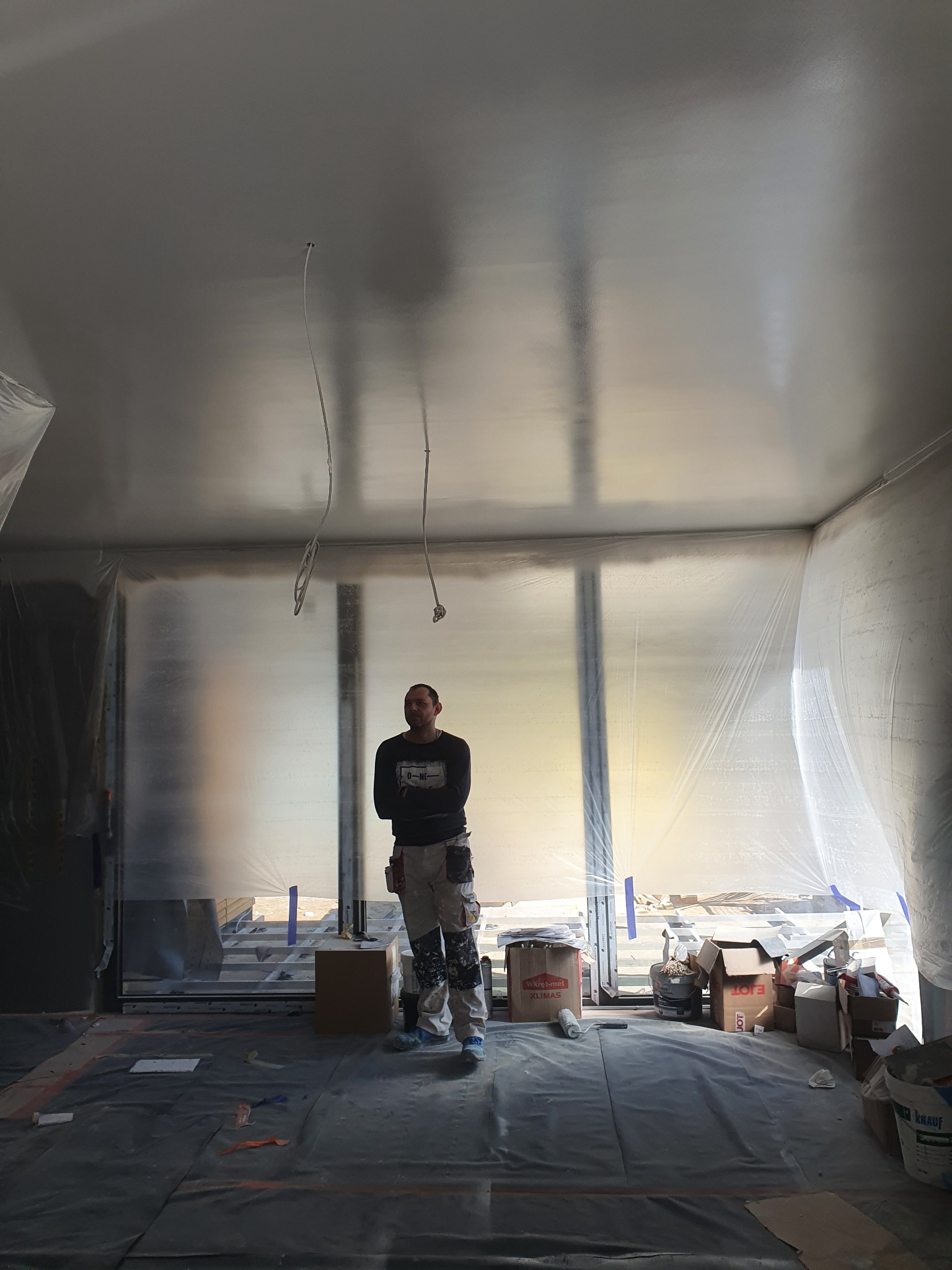 Mężczyzna w ubraniu roboczym stoi w pomieszczeniu przygotowanym do malowania, ściany i sufit zabezpieczone folią, widoczne narzędzia i materiały malarskie.