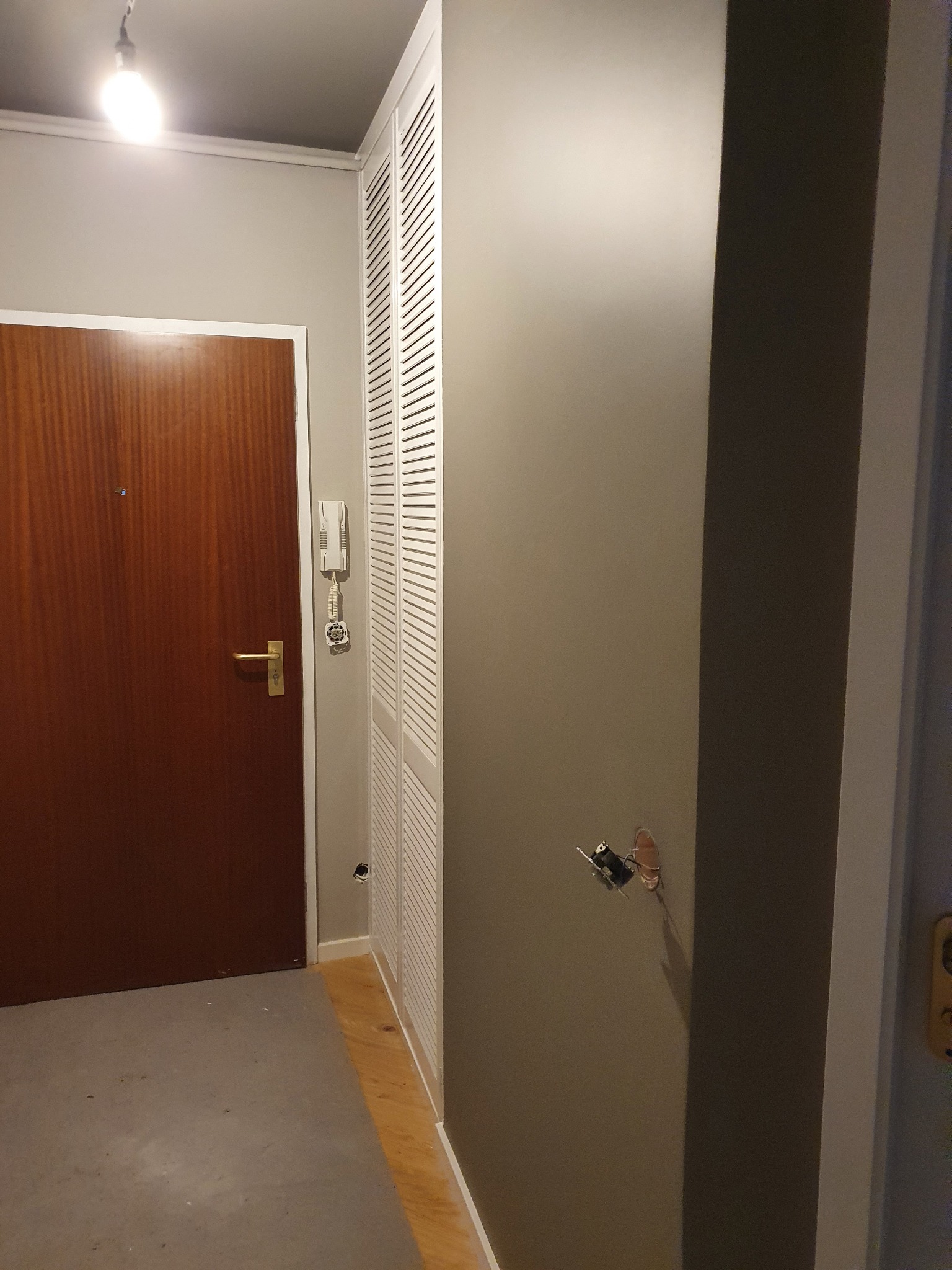 Korytarz po odświeżeniu koloru ścian na szaro, widoczne gniazdko elektryczne bez osłony, drewniane drzwi wejściowe i biała szafa z lamelami.