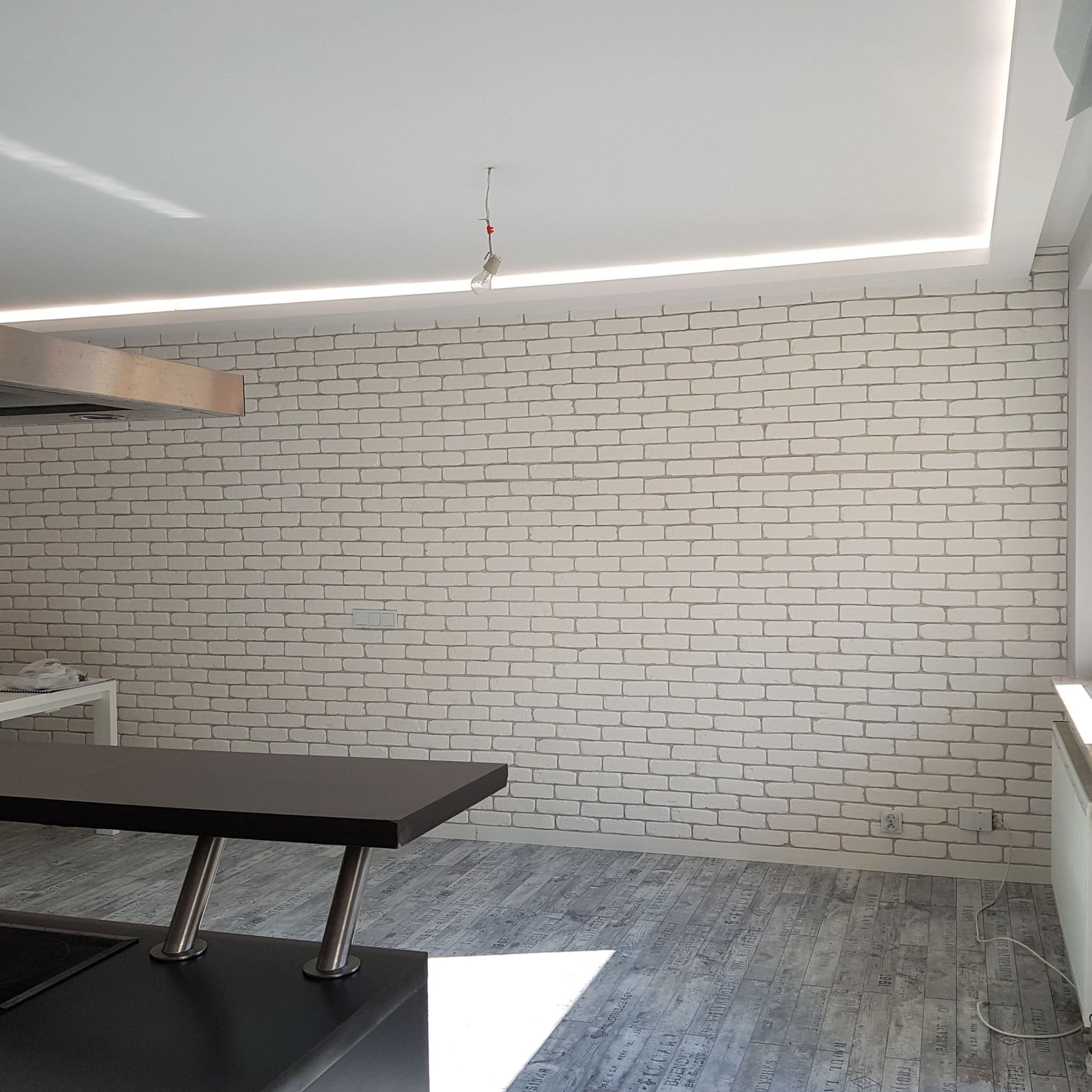 Kuchnia z białą ceglaną ścianą, podwieszanym sufitem z oświetleniem LED i niedokończonym podłączeniem lampy wiszącej.