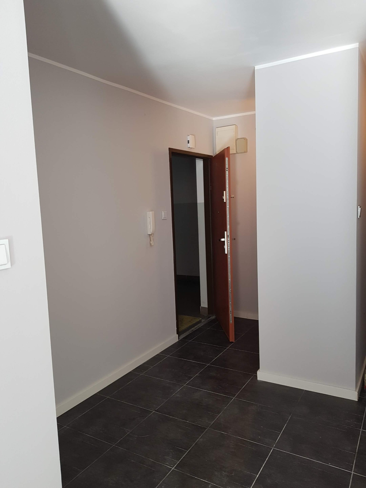 Wyremontowany korytarz z ciemną, kwadratową podłogą z płytek, świeżo pomalowanymi ścianami w odcieniach szarości i otwartymi drzwiami wejściowymi.