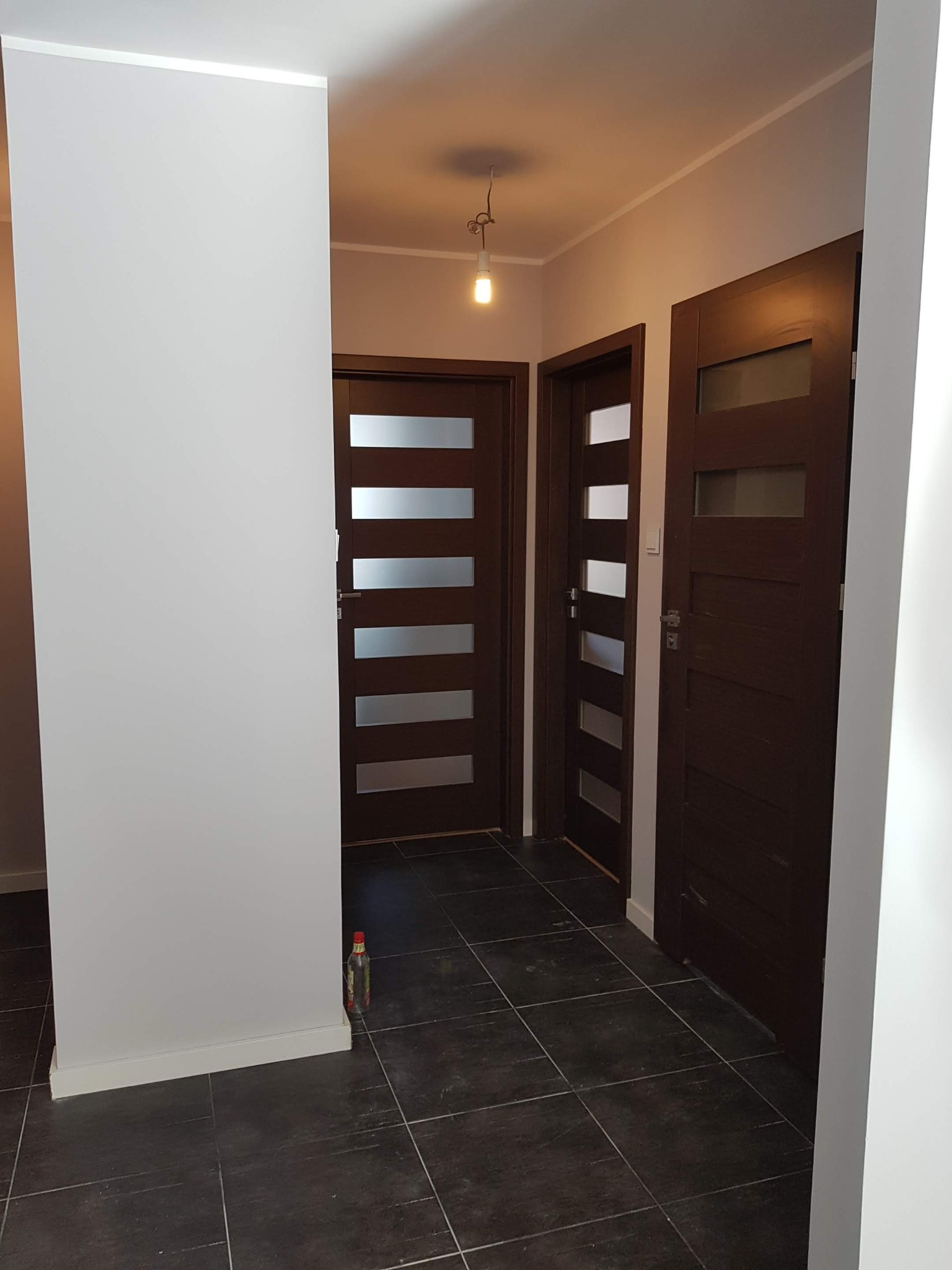 Wnętrze korytarza po odświeżeniu: szare ściany, ciemna podłoga z płytek i trzy ciemnobrązowe drzwi z poziomymi przeszkleniami. Na podłodze stoi butelka z płynem.