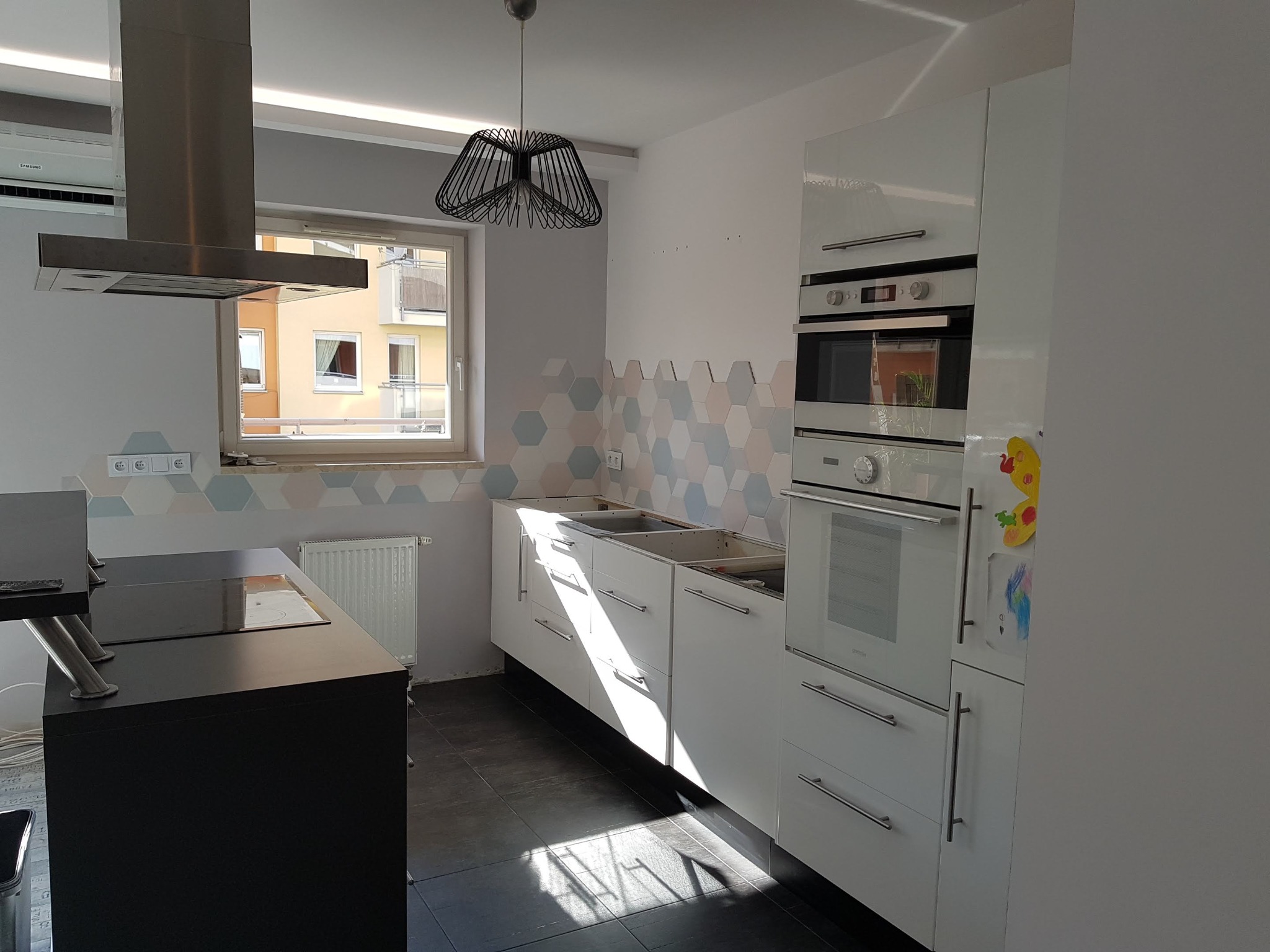 Wnętrze kuchni w trakcie montażu: białe szafki bez blatów, piekarnik w zabudowie, heksagonalne płytki na ścianie, czarna wyspa kuchenna, nowoczesna lampa wisząca.