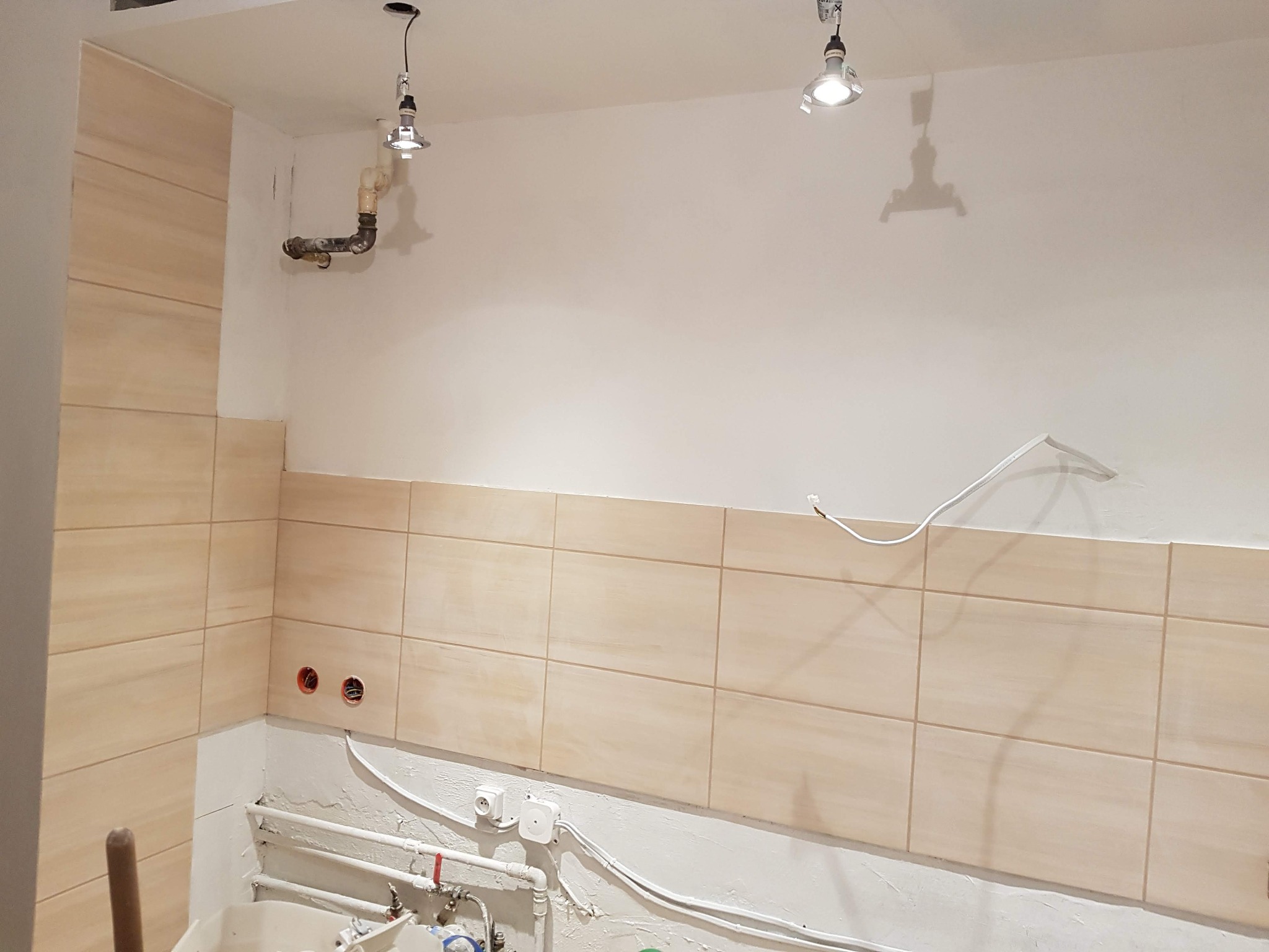 Łazienka w trakcie prac wykończeniowych: ułożone beżowe płytki na ścianach, widoczne wystające przewody elektryczne, rury i niedokończone podłączenia hydrauliczne, surowe wykończenie sufitu...