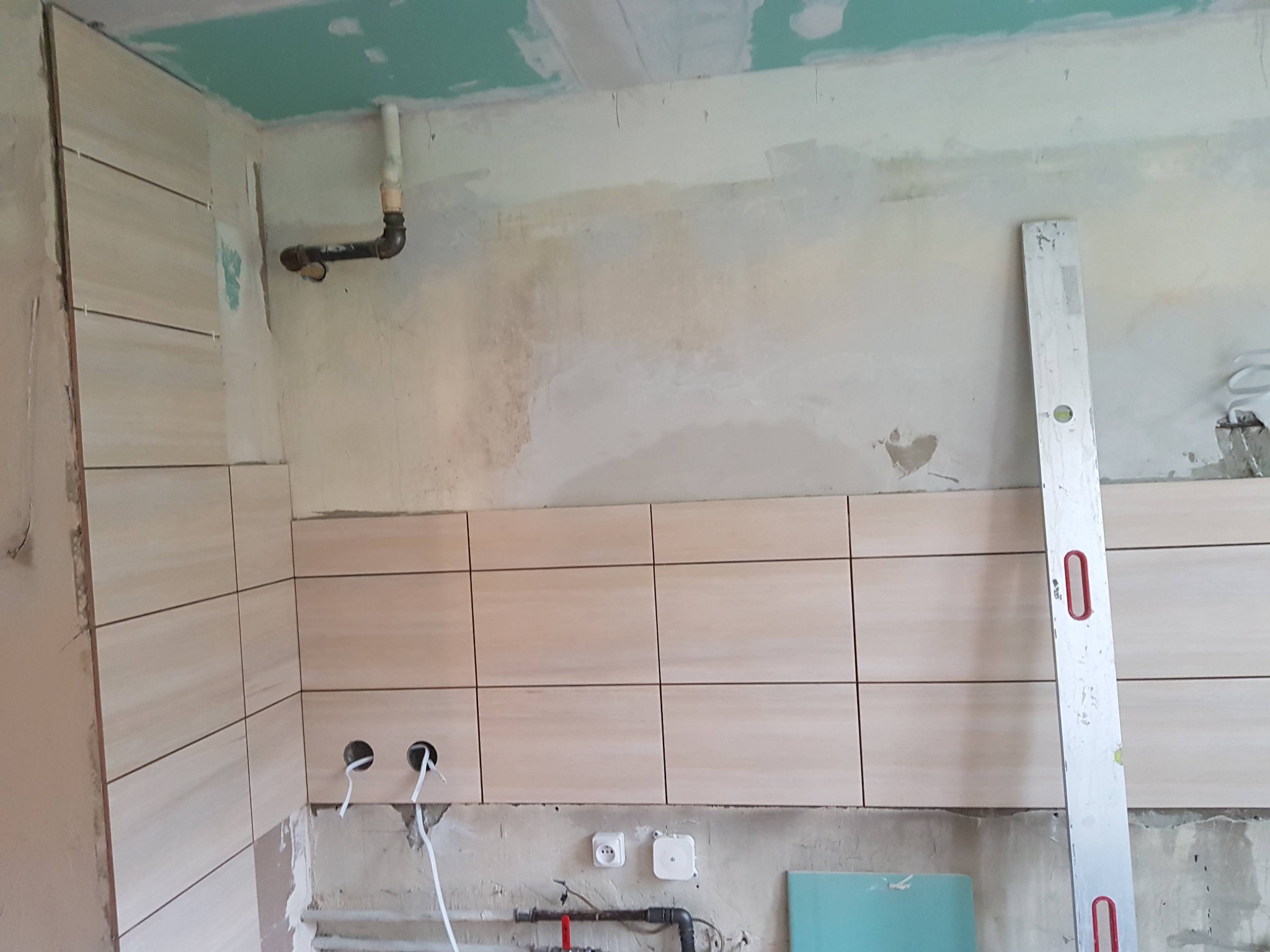 W trakcie kładzenia beżowych płytek na ścianie w łazience, widoczny poziomica przy ścianie, instalacja elektryczna i fragment sufitu z płyt kartonowo-gipsowych.