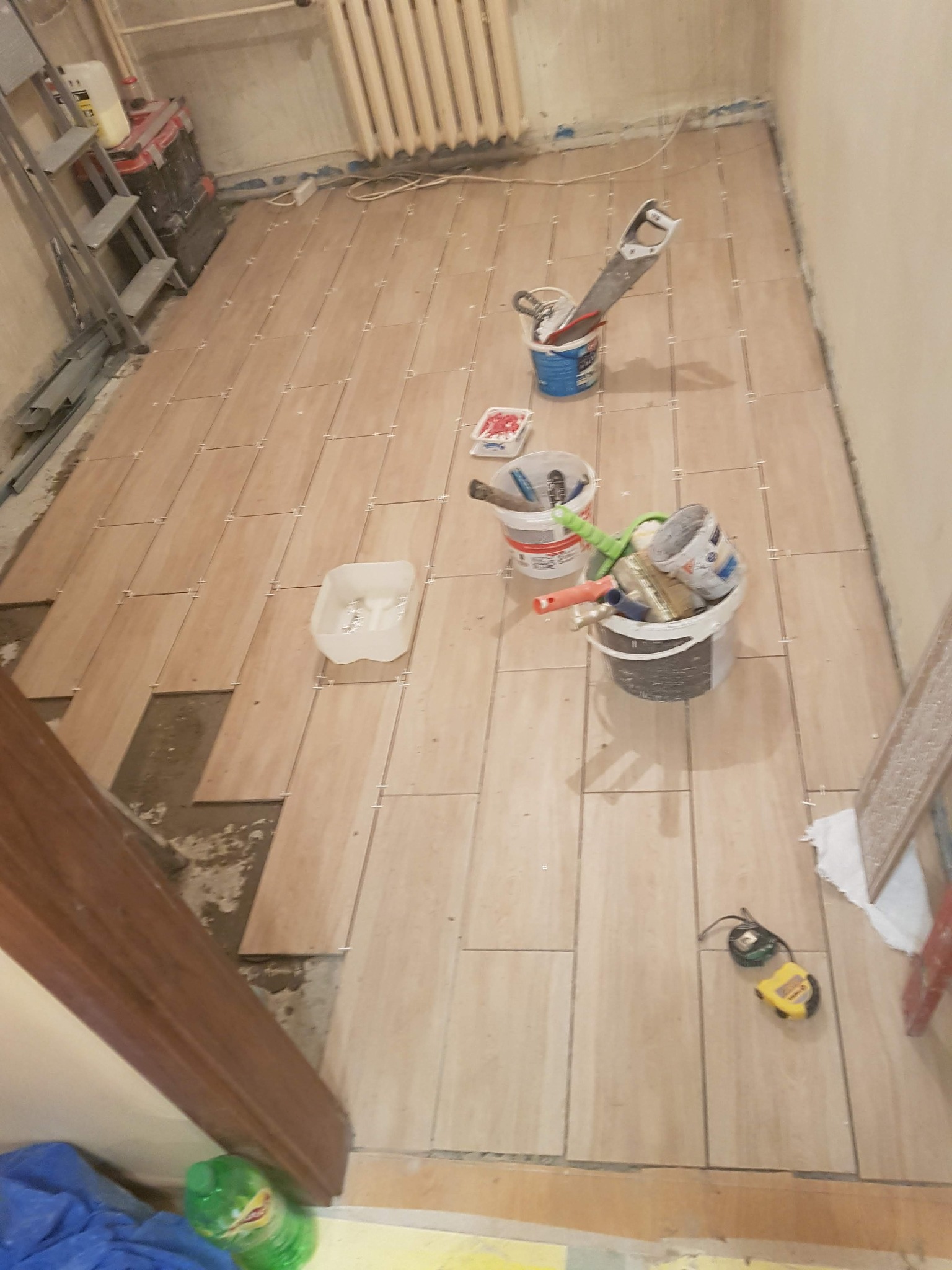Proces układania podłogi z płytek imitujących drewno, widoczne narzędzia: poziomica, miarka, wiadro z farbą, szczotki, szpachelki i krzyżyki dystansowe, częściowo ułożona podłoga z pozostawionymi...