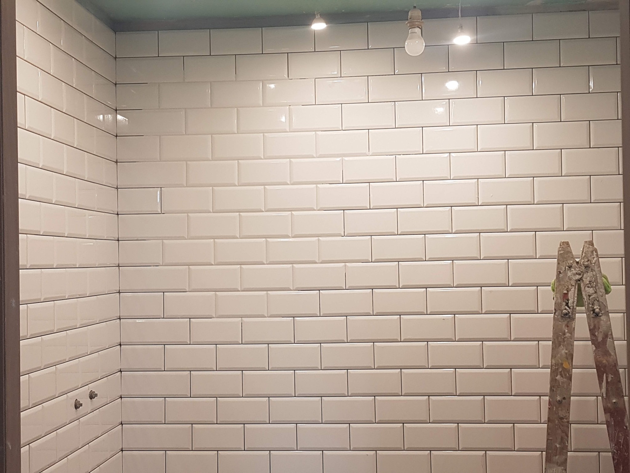 Ściana w łazience wyłożona białymi, błyszczącymi płytkami typu 'metro', z widoczną drabinką malarską z resztkami farby i prowizorycznym oświetleniem sufitowym.