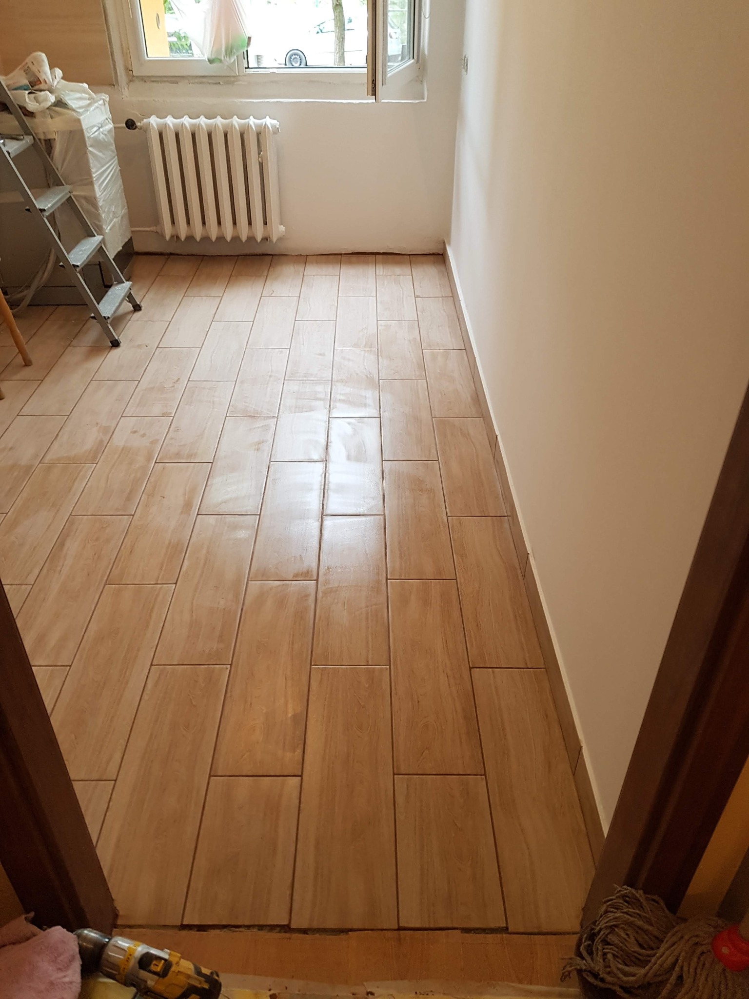 Ułożone podłużne płytki podłogowe imitujące drewno w jasnym pomieszczeniu z białymi ścianami i grzejnikiem, widok zza progu, drabina aluminiowa w tle.