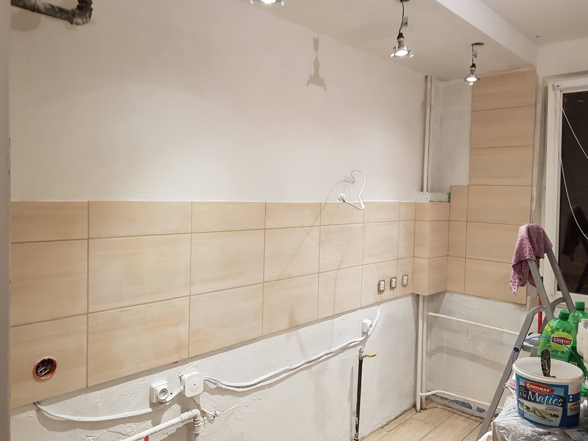 Układanie beżowych płytek w kuchni, widoczne puszki elektryczne, rury i drabina z farbą, etap prac wykończeniowych.