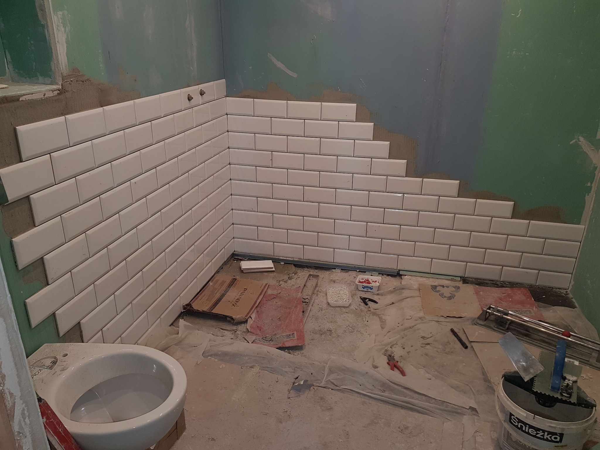 Montaż białych, prostokątnych płytek na ścianie łazienki w trakcie remontu, widoczne narzędzia glazurnicze i niepodłączona toaleta.