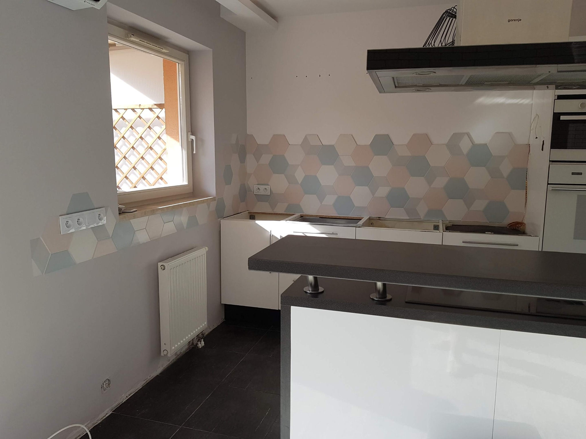 Wnętrze kuchni w trakcie montażu z heksagonalnymi płytkami w pastelowych kolorach, widoczny fragment wyspy kuchennej z czarnym blatem i białymi frontami, okno z kratką oraz grzejnik na szarej ścianie.