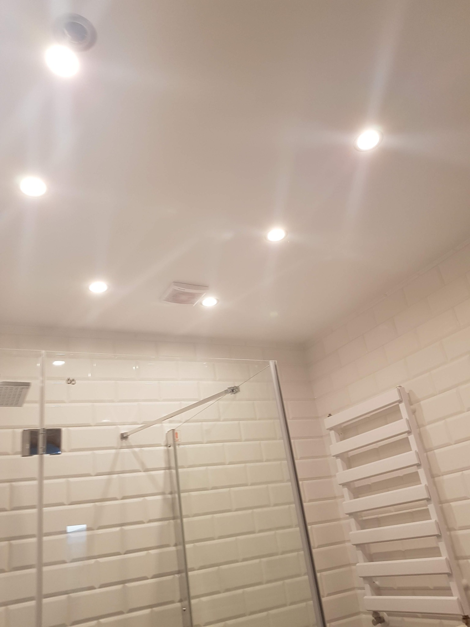 Nowoczesna łazienka z białymi, prostokątnymi płytkami imitującymi cegłę, szklaną kabiną prysznicową, grzejnikiem drabinkowym i wpuszczanymi lampami sufitowymi.