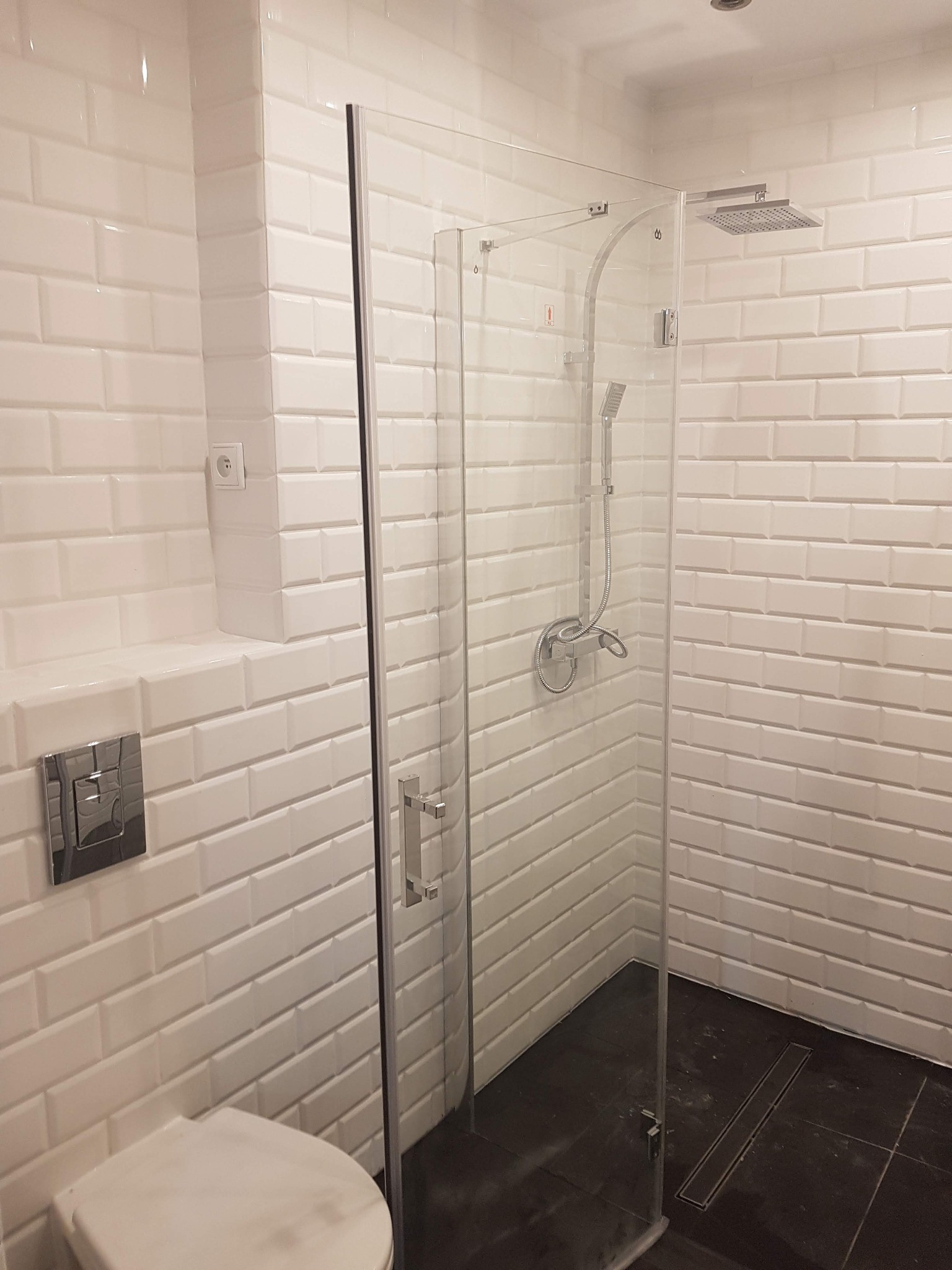 Nowoczesna łazienka z białą, cegiełkową glazurą, kabiną prysznicową ze szklanymi drzwiami i deszczownicą, oraz podwieszaną toaletą z chromowanym przyciskiem.