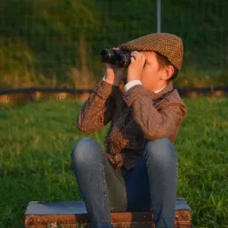 Chłopiec w brązowej kraciastej marynarce i kaszkiecie siedzi na walizce na trawie, patrząc przez lornetkę.