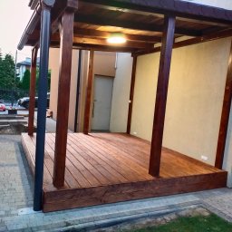 Artifex - Zadaszona konstrukcja z ciemnego drewna z oświetleniem punktowym na podłodze z desek, widok z zewnątrz na kostkę brukową i fragment ogrodu.