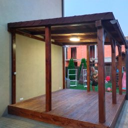 Artifex - Zadaszona konstrukcja z drewnianą podłogą, brązowe słupy, oświetlenie sufitowe, widoczna osoba w tle, plac zabaw i budowa w sąsiedztwie.