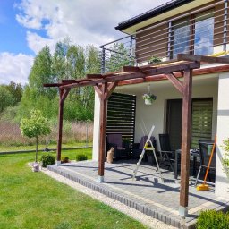 Artifex - Nowoczesna pergola z ciemnego drewna przylegająca do domu z balkonem, widok z trawnika w słoneczny dzień, drabina oparta o konstrukcję.