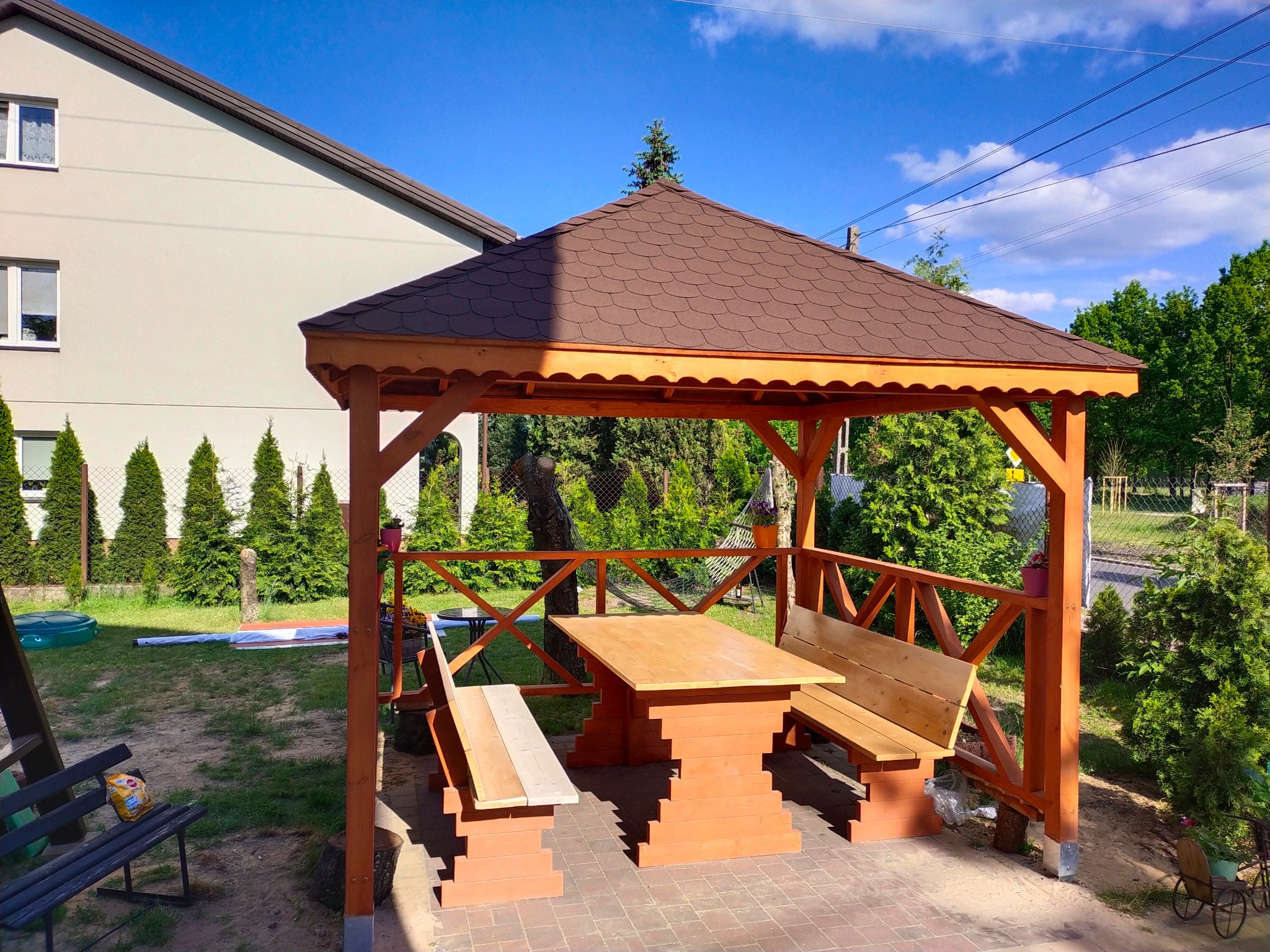 Nowo wybudowana, drewniana altana z brązowym dachem krytym gontem bitumicznym, wyposażona w stół i ławki z jasnego drewna, ustawiona na brukowanym podłożu w przydomowym ogrodzie.