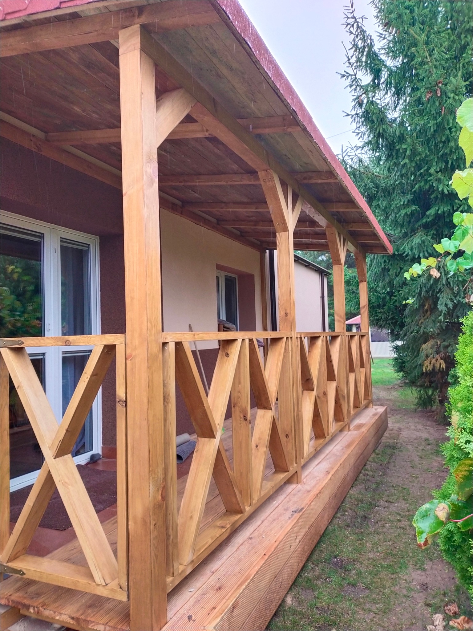 Zadaszona konstrukcja z drewnianą balustradą o geometrycznym wzorze, przylegająca do budynku z widocznymi oknami, otoczona zielenią w pochmurny dzień.