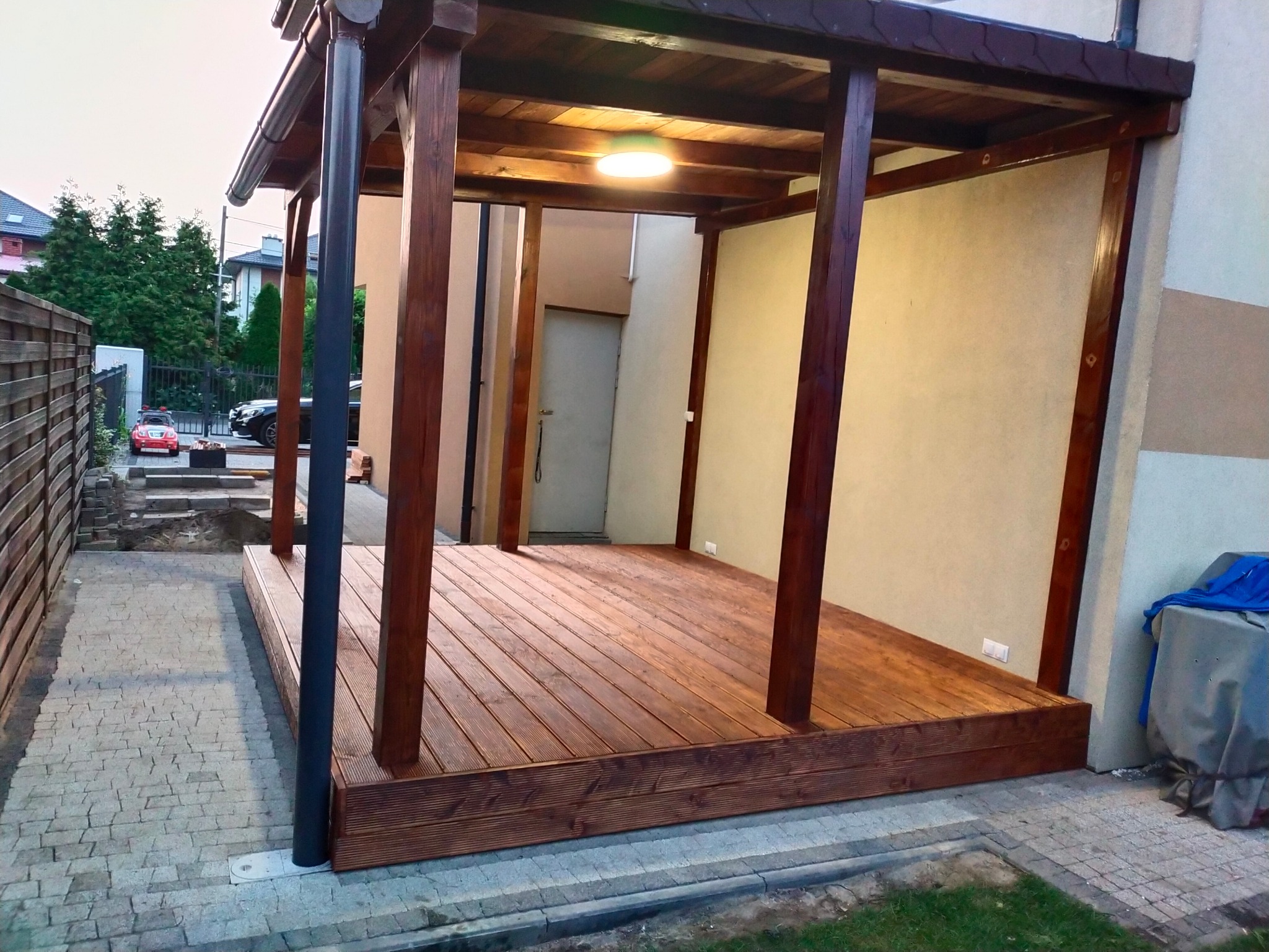 Zadaszona konstrukcja z ciemnego drewna z oświetleniem punktowym na podłodze z desek, widok z zewnątrz na kostkę brukową i fragment ogrodu.