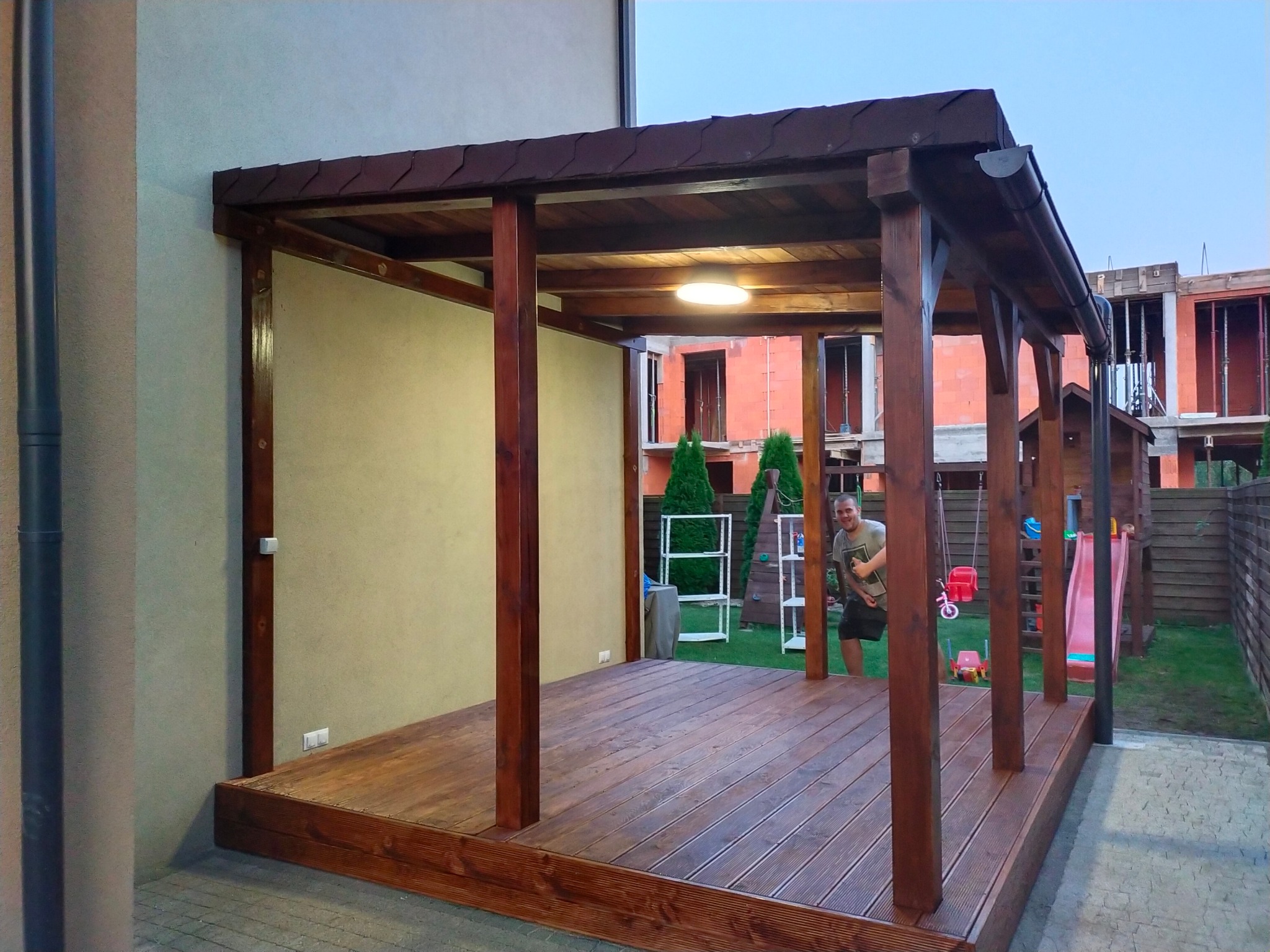 Zadaszona konstrukcja z drewnianą podłogą, brązowe słupy, oświetlenie sufitowe, widoczna osoba w tle, plac zabaw i budowa w sąsiedztwie.