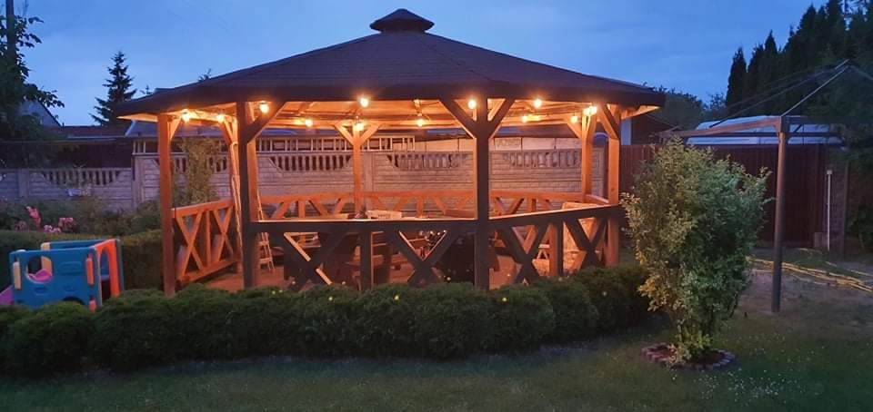 Drewniana altana z oświetleniem w ogrodzie, widoczny plac zabaw dla dzieci w tle.