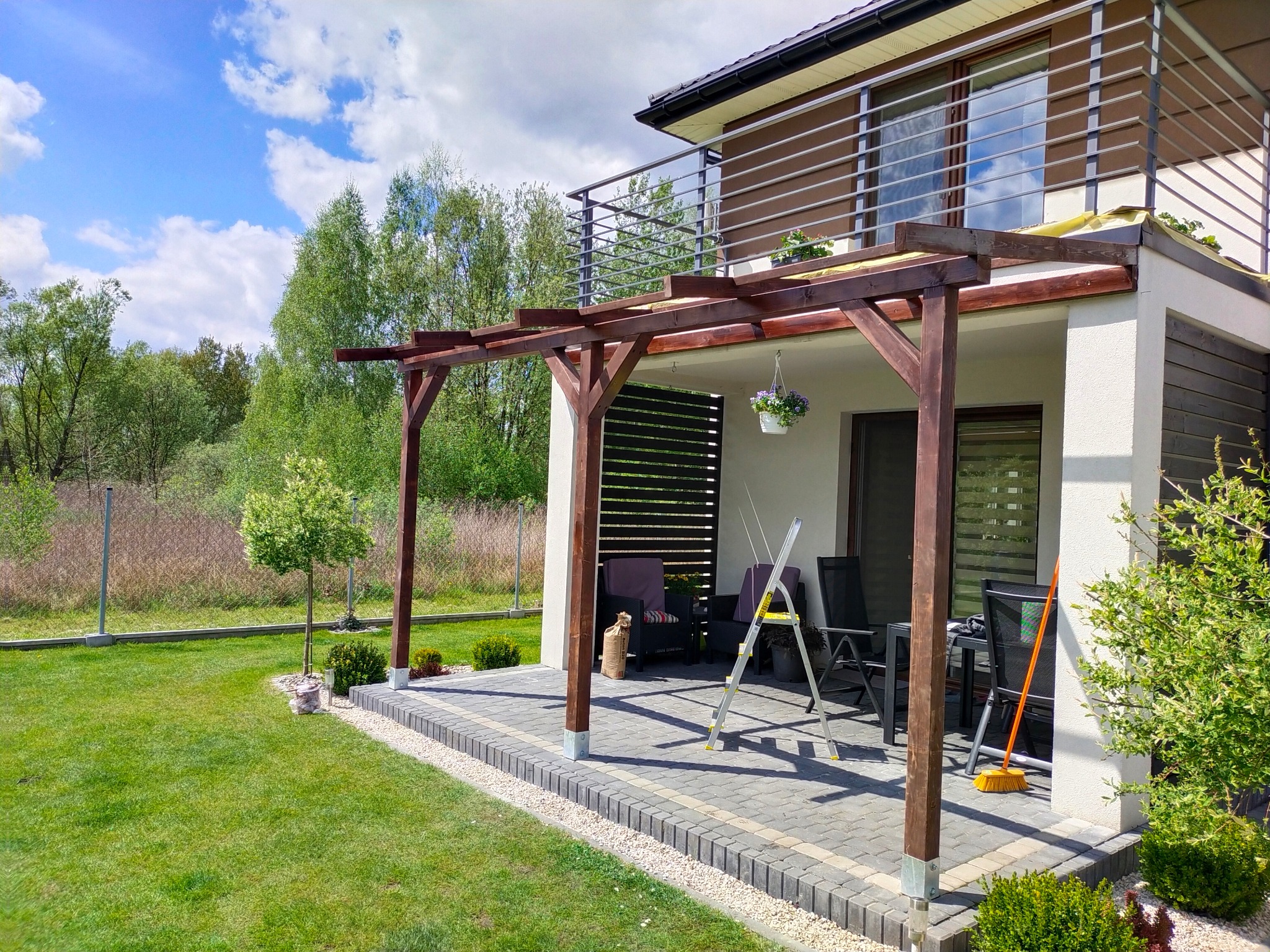 Nowoczesna pergola z ciemnego drewna przylegająca do domu z balkonem, widok z trawnika w słoneczny dzień, drabina oparta o konstrukcję.