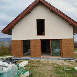 BOB BUD - Dwukondygnacyjny dom jednorodzinny z drewnopodobną elewacją w trakcie montażu, widoczne płyty styropianowe i puszki farb na trawniku.