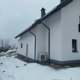 Biały dom z szarymi elementami wykończeniowymi i klimatyzatorem na zewnątrz, otoczony śniegiem w zimowy dzień. Widok z boku, z elementami placu budowy w tle.